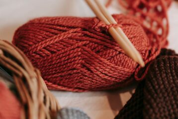 Skein of red yarn with wooden knitting needles placed on soft couch with crop faceless person in light room at home