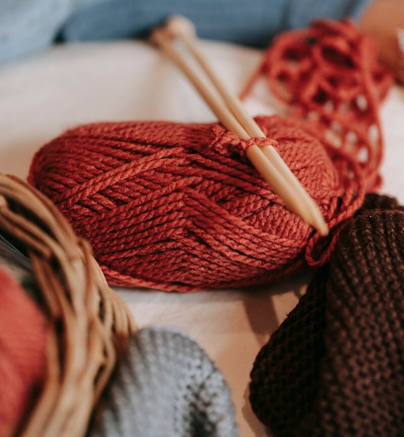 Skein of red yarn with wooden knitting needles placed on soft couch with crop faceless person in light room at home