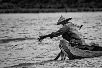 grayscale photo of man in hat and long sleeve shirt sitting on boat on water