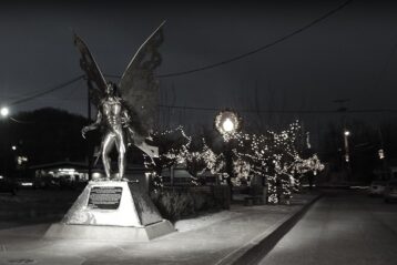 black and white photo of the mothman statue in point pleasant, west virginia, taken at night with christmas lights visible in the distance