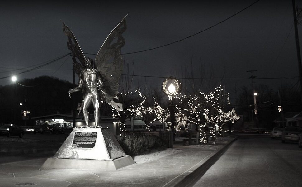 black and white photo of the mothman statue in point pleasant, west virginia, taken at night with christmas lights visible in the distance