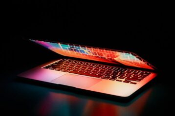 Laptop partially opened, emitting vibrant multicolored lights in dark setting.